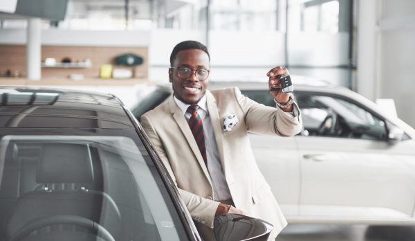 Handsome black man in dealership is hugging his new car and smiling.